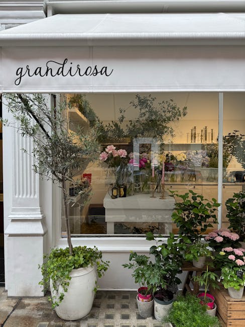 The image shows the exterior of a flower shop named 'Grandirosea,' with a large glass window displaying an array of vibrant floral arrangements indoors. In front of the shop, there are several potted 