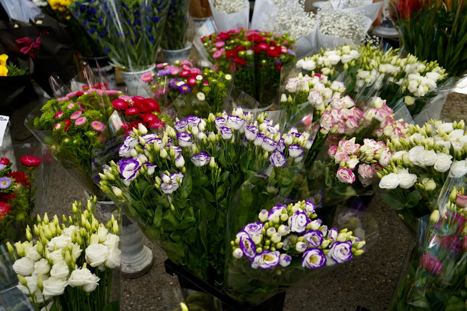 A variety of freshly cut flowers arranged in bouquets at a florist shop, including purple and white lisianthus, pink calendulas, white roses, and other colorful blossoms, all neatly wrapped in transpa