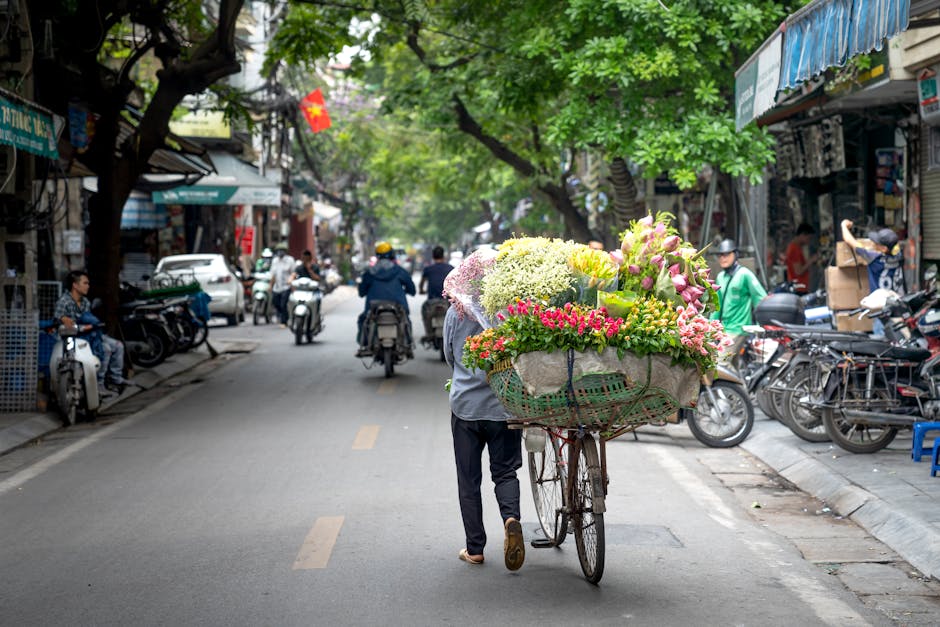 A street scene in an urban area with a person pushing a bicycle loaded with a large, colorful floral arrangement comprising pink, white, and green flowers, including lilies, roses, and hydrangeas, wra
