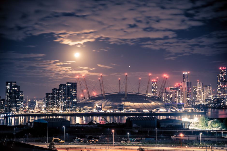 A nighttime cityscape featuring the illuminated London Stadium with its distinctive domed roof surrounded by modern high-rise buildings, some under construction with cranes and red warning lights. The