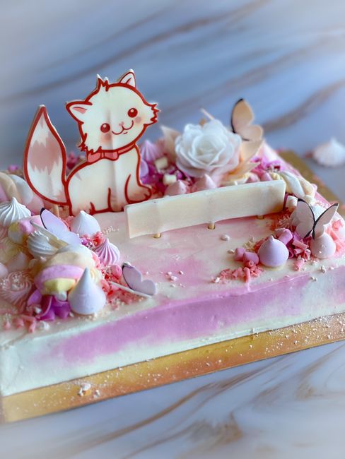 A rectangular cake decorated with pink and white buttercream frosting, featuring a cute white chocolate fox topper with pink accents, surrounded by small pastel meringues, pink and white sugar confect